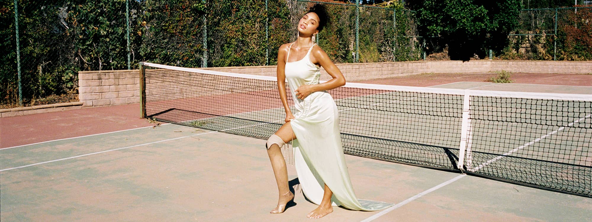 Model Lyric Mariah stands on a tennis court wearing a long silk green dress with her hand on her hip. Her hair is in an updo and her skin is gleaming in the sun. She is popping out her prosthetic leg.